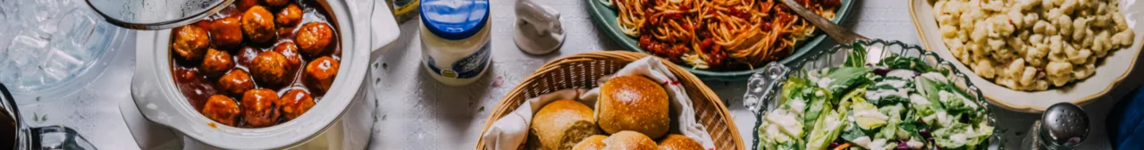 A potluck table with meatballs, dinner rolls, salad, spaghetti and a jar of Blue Plate mayonnaise