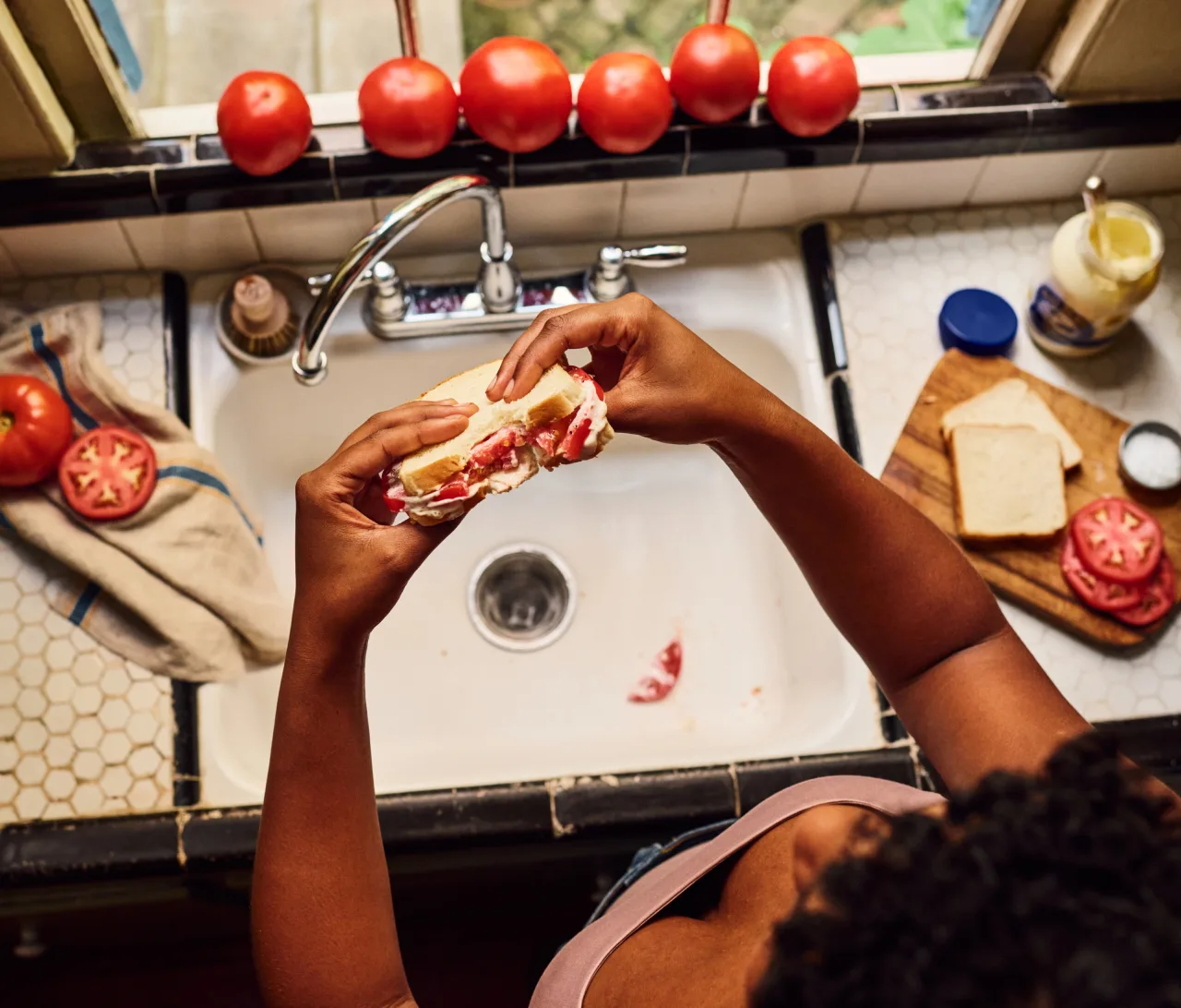 Hands holding a tomato sandwich over a kitchen sink